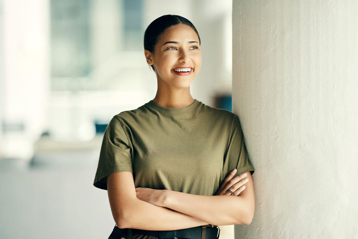 A smiling woman in an olive-green shirt standing against a light wall, representing the military families served by medical weight loss care at Optimal Well MD in Virginia Beach.