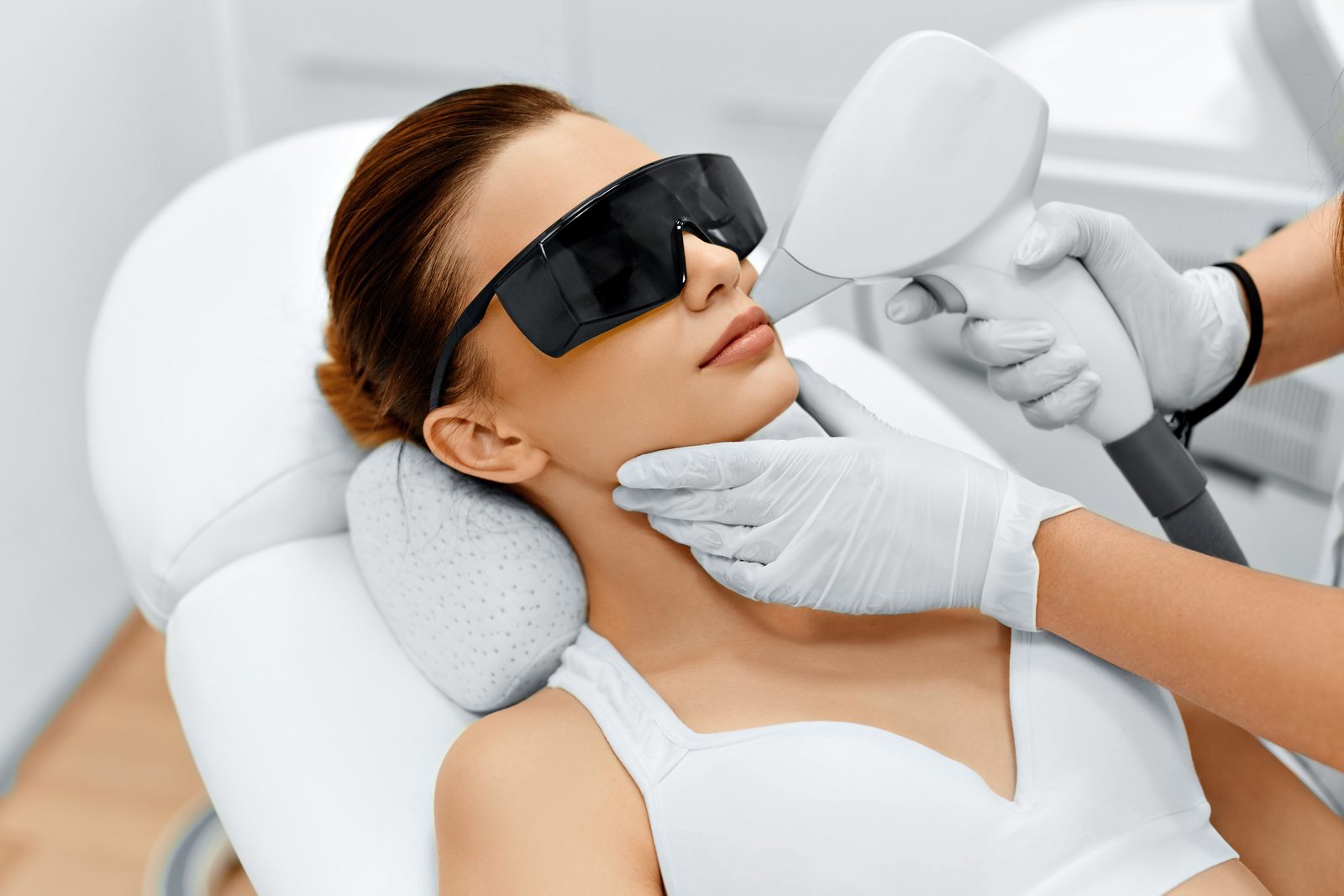 A clinician in gloves performing a laser hair removal treatment on a patient wearing protective eyewear in a clinical treatment room, illustrating laser hair removal costs in Virginia Beach