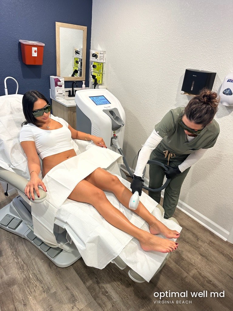 A patient reclines in a treatment chair wearing protective eyewear while a provider treats her leg with a laser hair removal device at Optimal Well MD in Virginia Beach