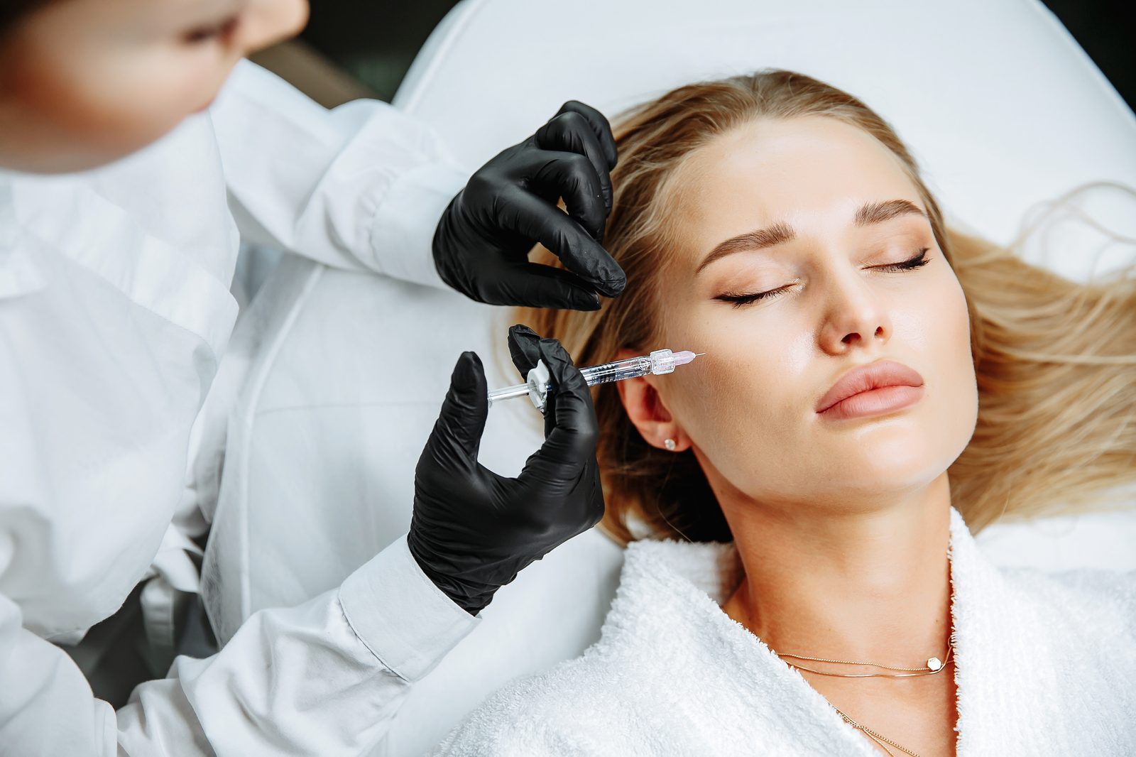 A woman reclining in a treatment chair while a gloved provider injects the mid-cheek area, representing Sculptra collagen-stimulating treatment in Virginia Beach
