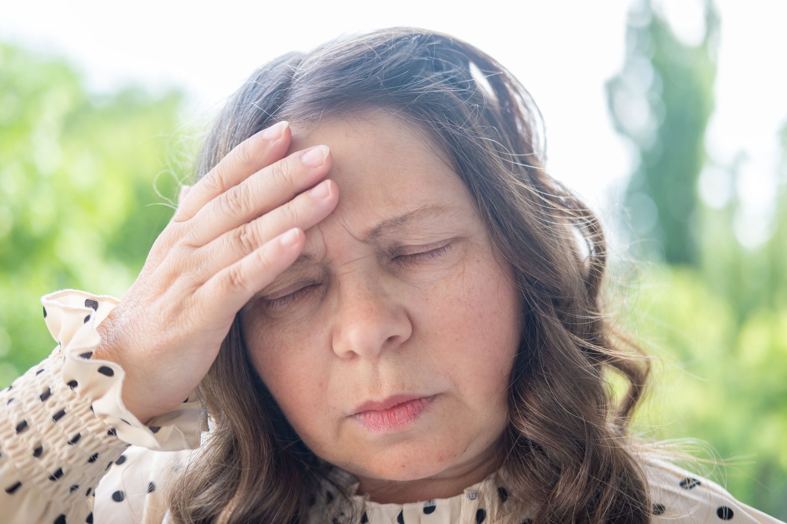A woman in her fifties outdoors resting a hand against her forehead during a hot flash, illustrating symptoms of hormone imbalance evaluated at a Virginia Beach wellness practice